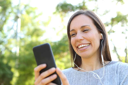 Happy adult woman listening to music checking smart phone online content in a park