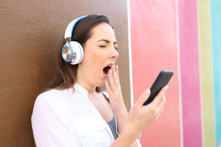 Bored woman yawning using smart phone and headphones to listen to music in a colorful street