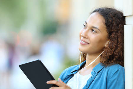 Happy mixed race woman dreaming looking at side listening to music with tablet and earphones in the streetの写真素材