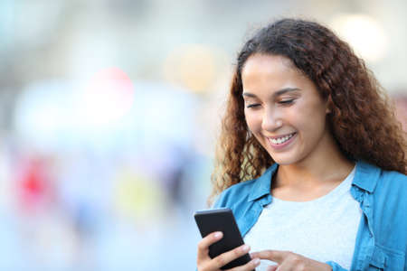 Happy mixed race woman using smart phone walking in the streetの写真素材