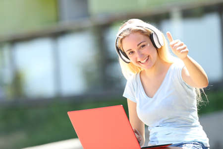 Happy student e-learning gesturing thumbs up sitting outdoors in an university campusの写真素材