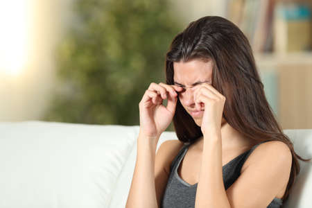 Girl suffering itching scratching eyes sitting on a couch in the living room at homeの写真素材