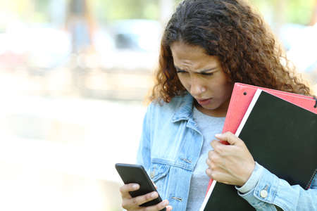 Sad mixed race student checking smart phone messages standing in a park or campusの写真素材