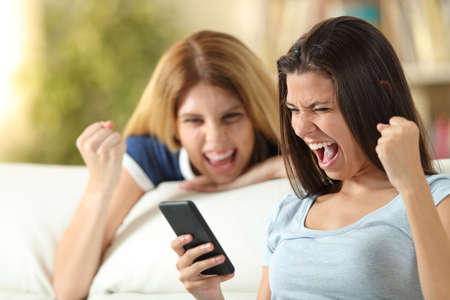 Two excited friends checking smart phone news sitting on a couch in the living room at homeの写真素材