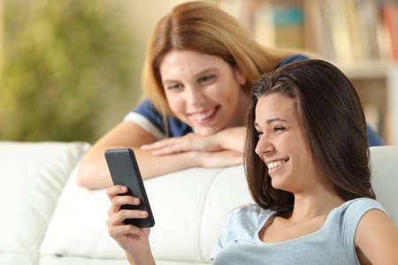 Two happy friends watching smart phone content sitting on a couch in the living room at homeの写真素材