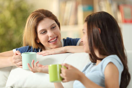 Happy friends talking at home drinking coffee sitting on a couch in the living room at homeの写真素材