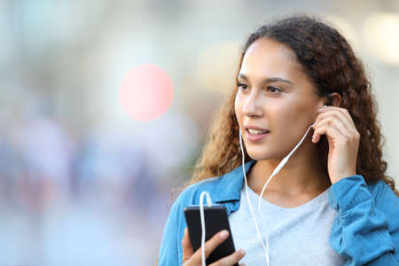 Mixed race woman holding smart phone putting earphones listening to music looking at side walking in the streetの写真素材