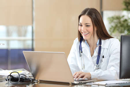 Happy doctor using a laptop writing content at consultationの写真素材