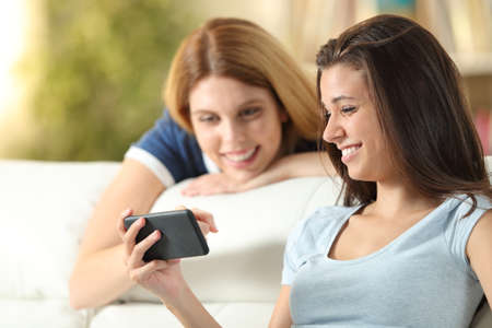 Two happy friends watching media content on phone sitting on a couch in the living room at homeの写真素材