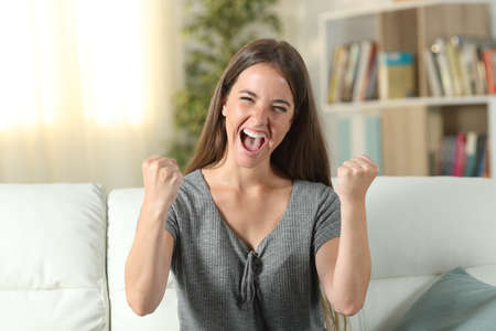 Front view portrait of an excited woman looking at camera sitting on a couch at homeの写真素材