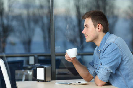 Serious man looking outdoors through a window holding a cup in a coffee shop in a winter eveningの写真素材