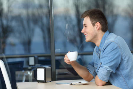 Happy man looking outdoors through a window holding a cup in a coffee shop in a winter eveningの写真素材