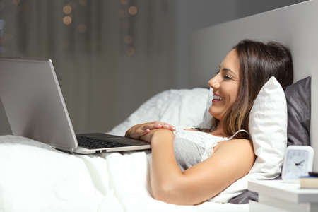 Happy woman uses a laptop watching videos lying on the bed in the night at homeの写真素材
