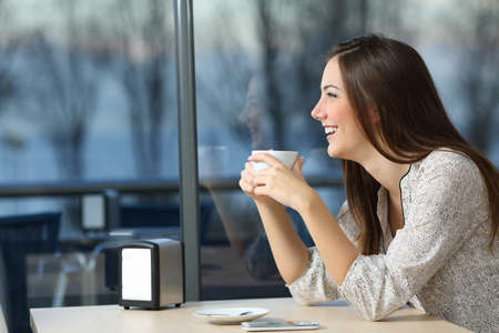 Happy girl looking outdoors through a window holding a cup in a coffee shop in a winter eveningの写真素材