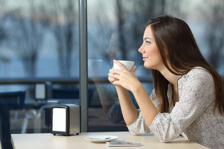 Serious girl looking outdoors through a window holding a cup in a coffee shop in a winter eveningの写真素材