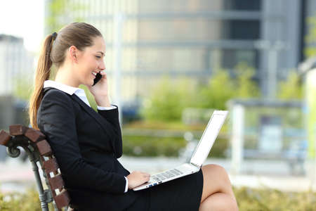 Side view portrait of a happy businesswoman talking on phone and checking laptop sitting on a benchの写真素材