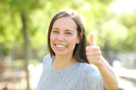 Happy woman looks at camera with thumbs up standing outdoors in a parkの写真素材