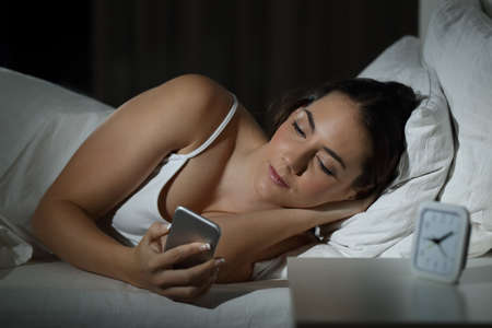 Serious woman holding smart phone checking content on the bed in the night at homeの写真素材