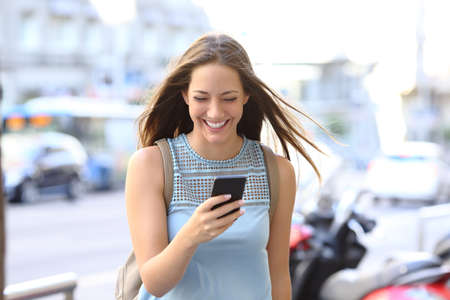 Front view portrait of a happy woman laughing walking towards camera  watching smart phone content in the streetの写真素材