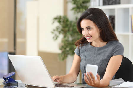 Happy office worker working on line using smart phone and laptop on a deskの写真素材