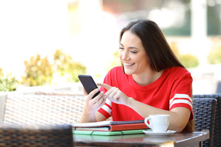 Happy student in red browsing phone content sitting in a coffee shop terraceの写真素材