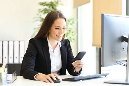 Happy businesswoman checking smart phone text sitting at officeの写真素材