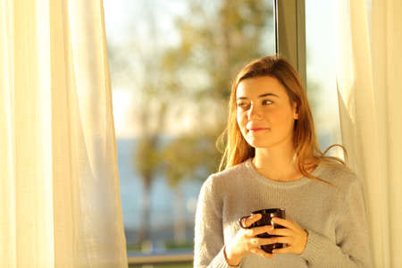 Happy teenage girl holding a hot drink thinking at home at sunsetの写真素材