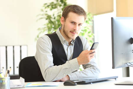 Serious executive holding smart phone looking at camera sitting at officeの写真素材
