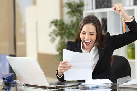 Excited businesswoman reading good news in letter sitting at officeの写真素材