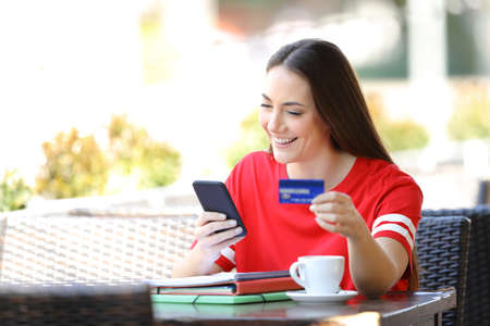 Happy student in red paying online with credit card sitting in a coffee shop terraceの写真素材