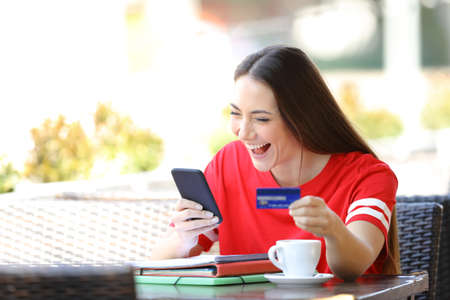 Excited student in red buying online sitting in a coffee shop terraceの写真素材