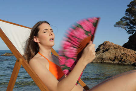 Unhappy sunbather suffering heat stroke fanning sitting on a deck chair on the beach on summer vacationの写真素材
