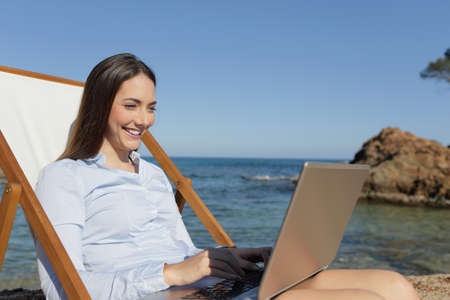 Happy entrepreneur using laptop sitting on a deck chair on vacation on the beachの写真素材
