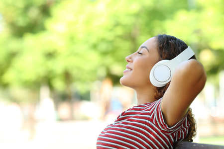 Side view portrait of a happy mixed race woman listening to music resting sitting on a bench in a parkの写真素材
