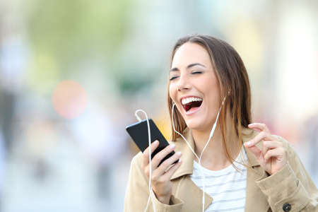 Funny girl wearing earphones listening to music and singing holding smart phone in the streetの写真素材
