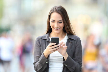 Front view portrait of happy woman using smart phone walking towards camera in the streetの写真素材