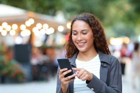 Happy mixed race woman using smart phone walking in the street with city lights in the backgroundの写真素材
