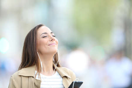 Satisfied woman wearing earphones listening to music on smart phone breathing fresh air in the streetの写真素材