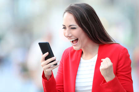 Excited lady in red celebrating good news checking smart phone on line content in the streetの写真素材