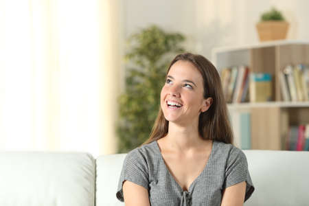 Front view portrait of a happy woman dreaming looking at side sitting on a couch at homeの写真素材