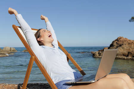 Excited entrepreneur with laptop raising arms celebrating success sitting on a deck chair on summer vacation on the beachの写真素材