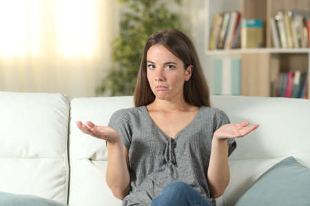 Front view portrait of a doubtful woman looking at camera sitting on a couch in the living room at homeの写真素材