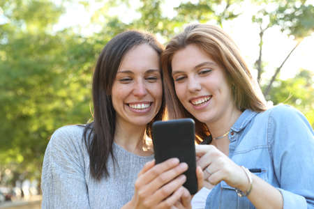 Front view portrait of an adult woman and teenage girl checking smart phone together in a parkの写真素材