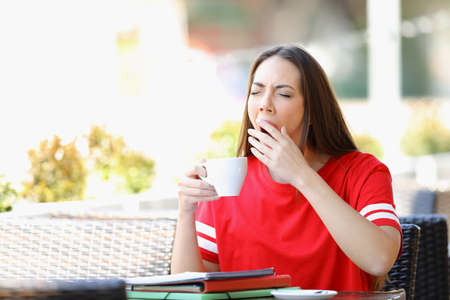 Tired student yawning in a bar holding a coffee cup sitting in a bar terraceの写真素材