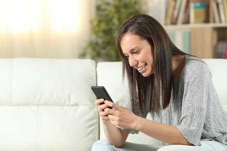 Happy woman using a smart phone sitting on a couch in the living room at homeの写真素材