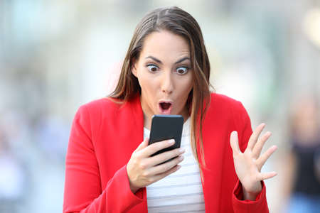 Surprised woman in red checking smart phone content standing in the streetの写真素材