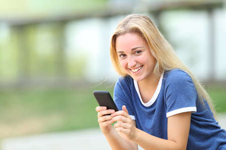 Teenage girl looking at camera holding smart phone sitting in an university campusの写真素材