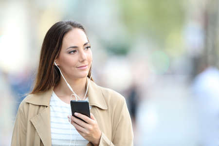 Front view portrait of a pensive woman wearing earbuds walking listening to music on phone in the streetの写真素材