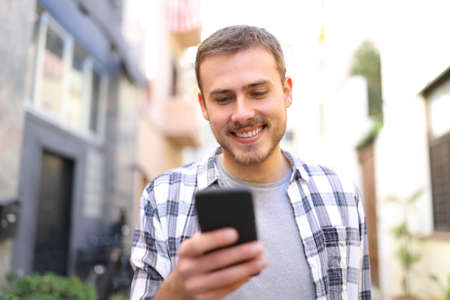 Front view portrait of a happy guy using smart phone walking towards you in the streetの写真素材