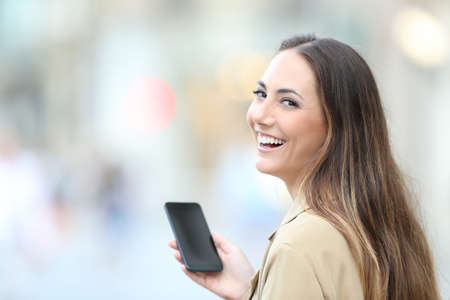Happy woman holding smart phone looks at camera standing in the streetの写真素材
